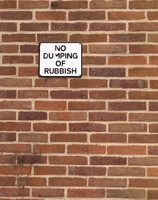 A rectangular metal sign mounted on a red brick wall displaying the message 'NO DUMPING OF RUBBISH' in black uppercase letters on a white background with a black border. The wall consists of uniformly laid bricks in shades of reddish-brown and dark brown, with light-colored mortar joints. The sign is positioned slightly above the center of the image, and the environment appears to be an outdoor urban or residential area. The brick wall's surface is slightly textured, and the overall scene suggests a controlled environment where waste disposal is regulated, aligning with the context of independent rubbish removal or private waste management services such as those offered by Rubbish Collection Wimbledon.