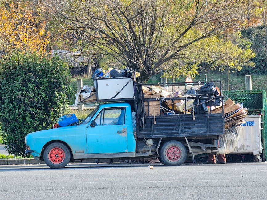 A small, vintage blue pickup truck parked on the side of a paved street, loaded with a large amount of waste materials including cardboard boxes, black plastic bags, and miscellaneous debris. The cargo extends above the truck bed and is secured with ropes, with sometimes loose items hanging or leaning over the sides. The truck's body shows signs of wear, with some fading paint and minor rust, especially around the wheel wells, and red-painted wheels that contrast with the vehicle's blue paint. The background features a lush green hedge, a tall deciduous tree with sparse leaves in shades of green and yellow, indicating an autumn setting, and a wooden fence behind the foliage. The environment appears to be a residential or suburban area during daylight, with soft natural lighting highlighting the textures of the waste and the truck’s surface. The scene reflects an instance of private waste collection or on-site clearance facilitated by a rubbish removal service like Rubbish Collection Wimbledon, emphasizing alternative waste handling methods.