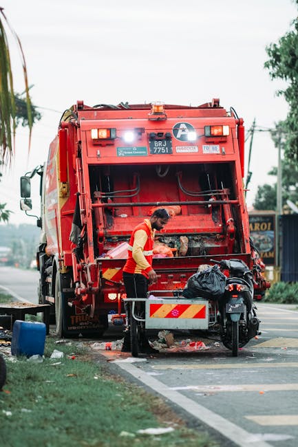 A street scene featuring a large red rubbish collection truck parked on the side of the road, with its rear compactor mechanism open and engaged in emptying waste. A worker wearing a high-visibility jacket and dark pants is positioned beside the truck, handling or inspecting the rubbish as it is being loaded. Behind the worker, a black plastic bag and other waste materials are visible near the truck's rear, with some debris scattered on the pavement. The environment includes a pathway with a curb and a grassy verge with a small blue bin or container on the left. In the background, there are trees, utility poles with wires, and a sign for a local business, indicating an urban or suburban setting. The scene is illuminated naturally, with overcast weather creating diffuse light, and the overall focus is on the waste collection process, demonstrating an aspect of private disposal services like those offered by Rubbish Collection Wimbledon, situated on a residential or commercial street.