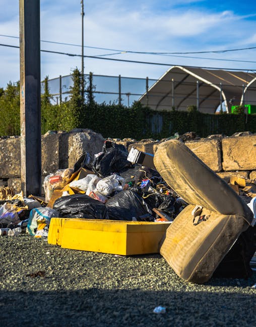 An outdoor scene featuring a pile of mixed waste materials on the ground, including black plastic garbage bags, a yellow plastic tray, and a discarded, worn tire leaning against the pile. The waste appears to consist of household refuse and debris, situated on a gravel surface. Behind the rubbish, there is a low stone wall made of roughly cut bricks, with lush green foliage and a tall metal fence visible beyond. Overhead, power lines cross the sky, which is partly cloudy with patches of blue. In the background, a structure with a curved, semi-transparent roof can be seen, possibly part of a sports facility or industrial building. The image reflects a typical scene where private waste collection services like Rubbish Collection Wimbledon might handle non-standard or excess rubbish that requires early removal or specialist disposal, especially outside regular municipal collection hours.