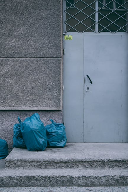 A street scene featuring a large red rubbish collection truck parked on the side of the road, with its rear compactor mechanism open and engaged in emptying waste. A worker wearing a high-visibility jacket and dark pants is positioned beside the truck, handling or inspecting the rubbish as it is being loaded. Behind the worker, a black plastic bag and other waste materials are visible near the truck's rear, with some debris scattered on the pavement. The environment includes a pathway with a curb and a grassy verge with a small blue bin or container on the left. In the background, there are trees, utility poles with wires, and a sign for a local business, indicating an urban or suburban setting. The scene is illuminated naturally, with overcast weather creating diffuse light, and the overall focus is on the waste collection process, demonstrating an aspect of private disposal services like those offered by Rubbish Collection Wimbledon, situated on a residential or commercial street.
