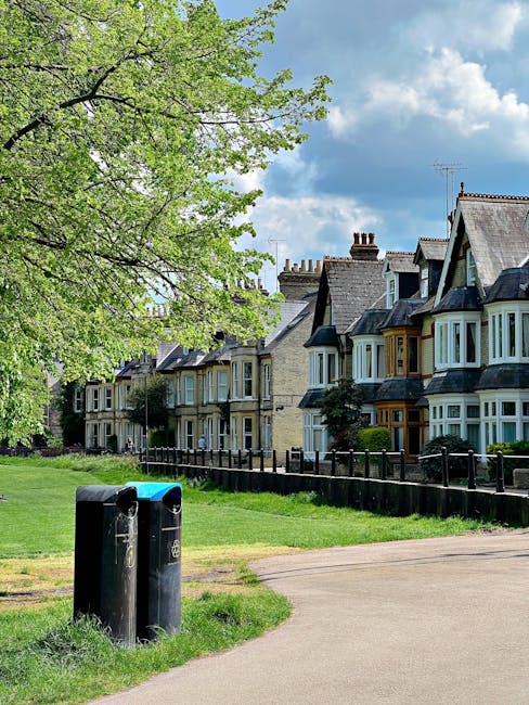 A street scene in a residential area featuring a row of Victorian-style terraced houses with bay windows, slate roofs, and brick chimneys. In the foreground, two black outdoor rubbish bins are positioned on a grassy verge next to a paved footpath. The bins have blue lids, indicating they are used for recycling or general waste, and are slightly weathered. To the left, a large deciduous tree with bright green foliage extends over the scene, creating partial shade. Behind the houses, a partly cloudy sky with patches of blue is visible. The environment appears calm and well-maintained, with a black metal fence running along the front of the houses, separating the gardens from the street, which aligns with private waste collection or alternative rubbish removal services like those offered by Rubbish Collection Wimbledon, particularly suited to narrow streets and urban settings.