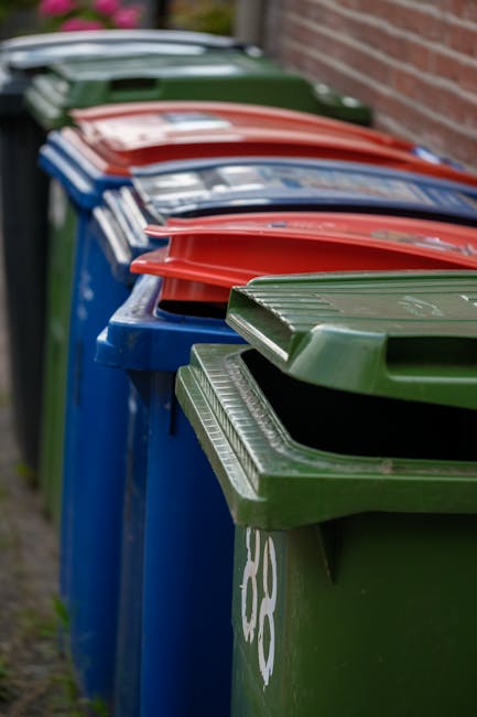 A row of standard wheelie bins lined up along a sidewalk beside a brick wall, each with hinged lids in different colours including green, red, blue, and grey. The green bin is positioned at the front, slightly to the right, with its lid partially open, revealing a dark interior. The blue and red bins are behind, with their lids closed, and the grey bin is further back, also with its lid shut. The bins are made of plastic, with textured surfaces and visible handles on the lids for opening. The background shows a blurred view of a paved pathway and some greenery, indicating an outdoor residential setting. The scene is evenly lit, with soft natural light highlighting the different textures and colours of the bins, which are likely used for separate waste or recycling collection services. The image relates to private waste management options in the context of alternative rubbish collection methods, as provided by Rubbish Collection Wimbledon.